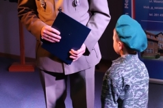 Żołnierz przemawia do mikrofonu. Obok niego stoi chłopiec w stroju żołnierza.