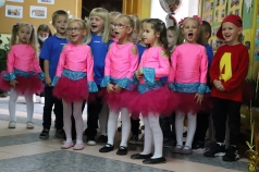 Na zdjęciu grupa śpiewających dziewczynek w różowych strojach. Chłopcy są w niebieskich koszulkach. Chłopiec stojący po prawej stronie na czerwoną bluzę z żółtą literą A i na głowie czerwoną czapkę z daszkiem