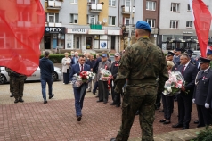 Na zdjęciu mężczyzna niosący kwiaty. Przed nim stoi żołnierz. Po prawej stronie ludzie ubrani na galowo