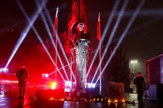 Pomnik „Anioła Wolności” oświetlony strumieniami światła. Po obu bokach stoi dwóch żołnierzy.
