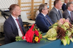 Na zdjęciu 4 mężczyzn siedzących za stołem. Na stole wiązanki kwiatów