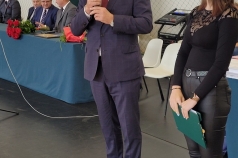 Na zdjęciu mężczyzna trzymający w ręku mikrofon. Obok niego uczennica. W tle mężczyźni siedzący za stołem