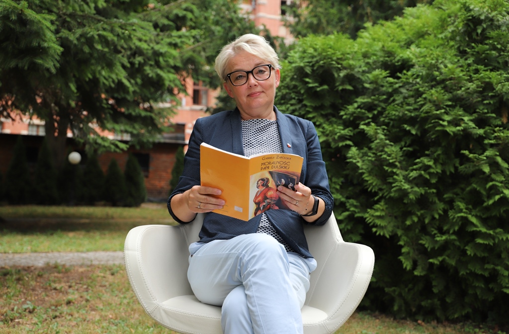 Na zdjęciu: w ogrodzie na białym krześle siedzi Zachodniopomorska Kurator Oświaty, w ręku trzyma książkę "Moralność Pani Dulskiej".