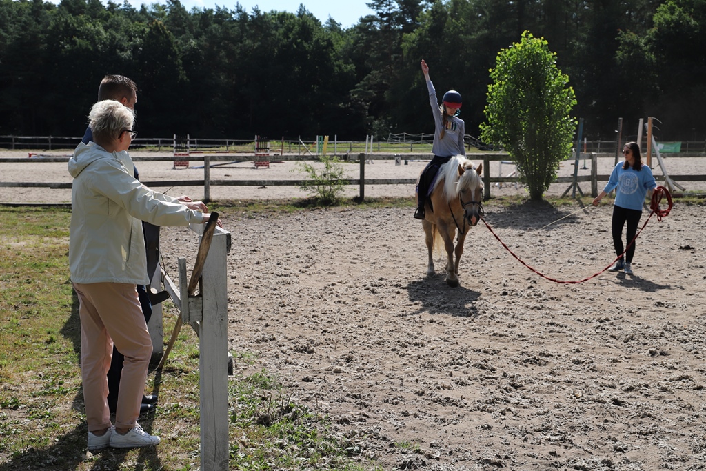 Na zdjęciu dziewczyna jeżdżąca na koniu. Obok stoi instruktorka. Po lewej stronie przyglądający się kobieta i mężczyzna