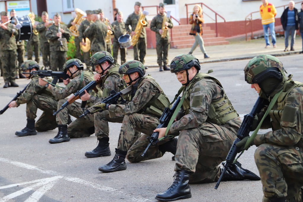 Grupa sześciu osób ubranych w strój żołnierza klęczy na ulicy i trzyma w ręku karabiny.