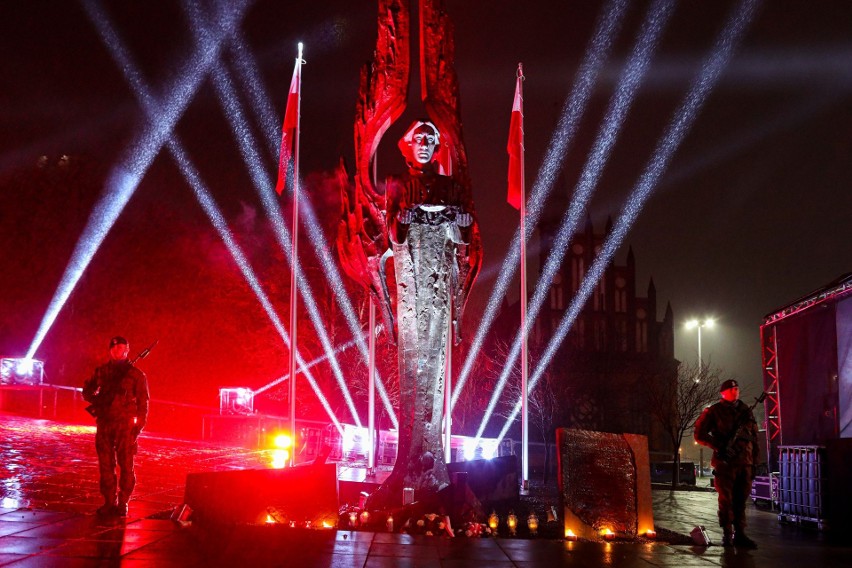 Pomnik „Anioła Wolności” oświetlony strumieniami światła. Po obu bokach stoi dwóch żołnierzy.