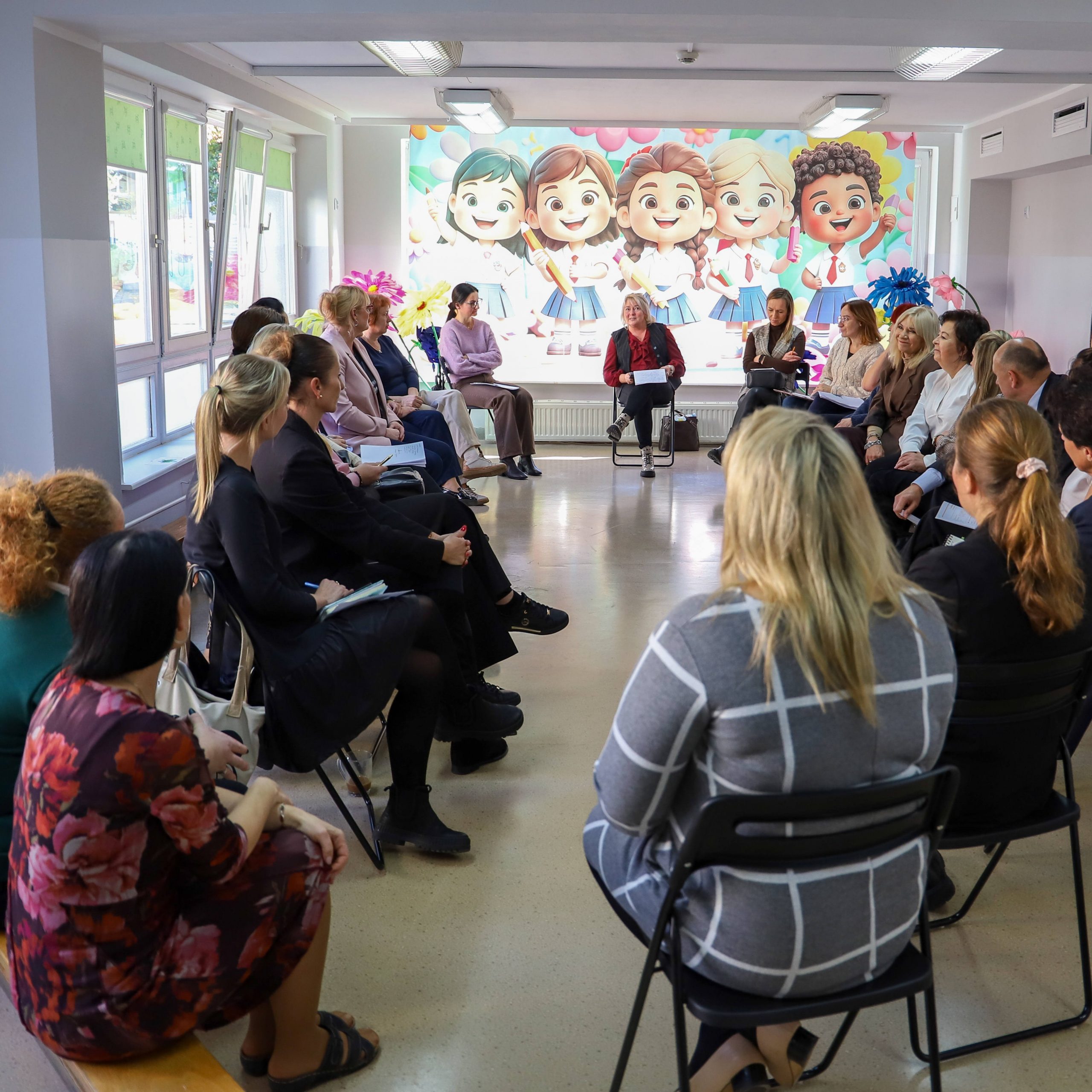 grupa dorosłych siedzi w okregu w tle kolorowy obrazem z dziećmi