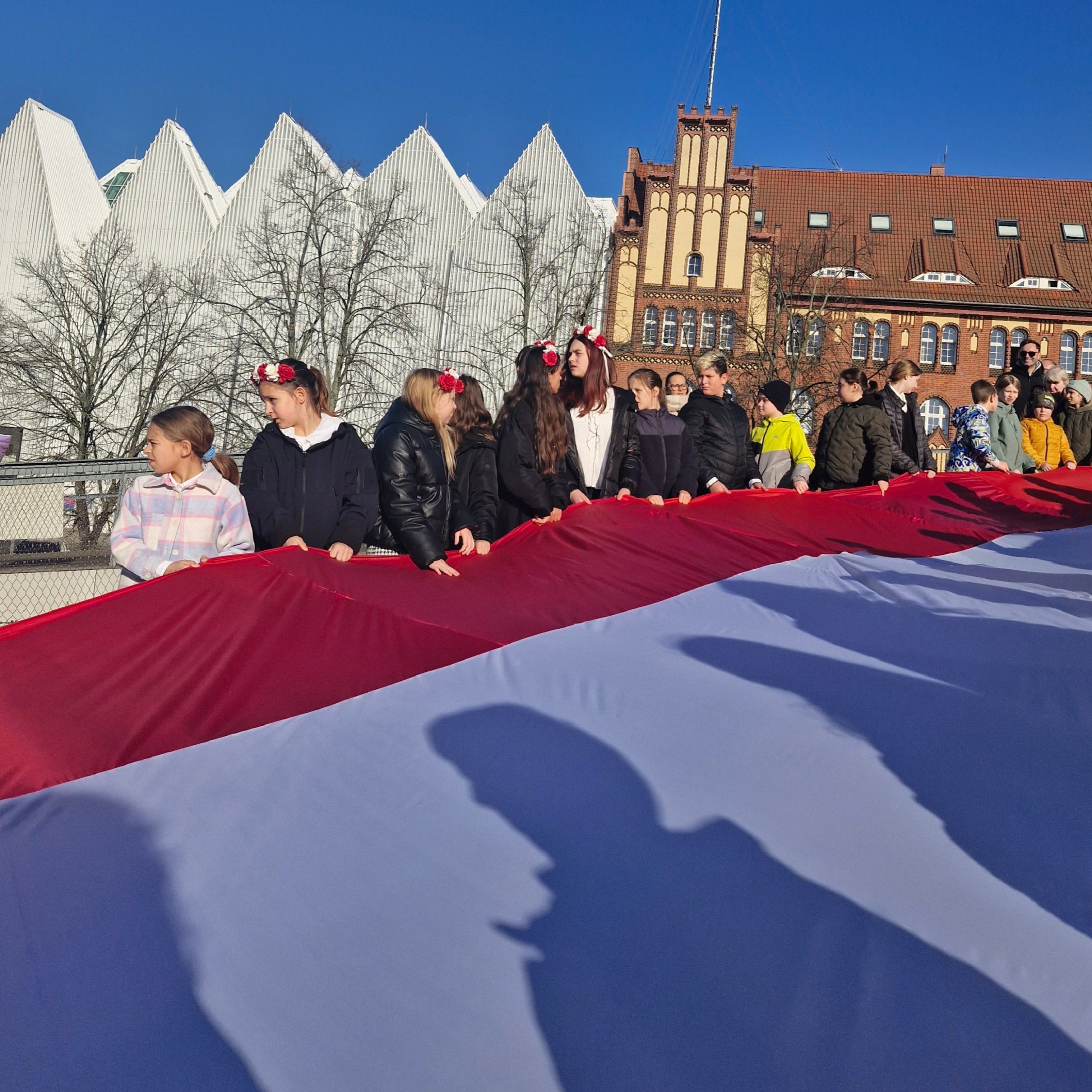 dzieci trzymają ogromną flagę biało-czerwoną, w tle filharmonia w Szczecinie i fragment Komendy Wojewódzkiej Policji przy placu Solidarności w Szczecinie